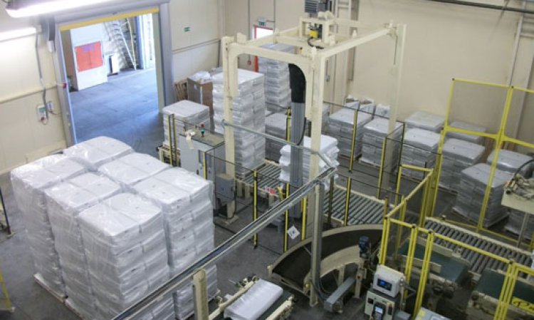 A bale (lower left) is conveyed to the model AR-200 series robotic palletiser (right) and onward to the model RA13/11 rotary arm stretch wrapper (left). A forklift that moves stretch-wrapped pallets through the doorway is the only manual step in this otherwise automated process.